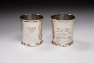 A color photograph of two engraved silver cups. One is embossed with an ornate "W" and the other reads "Camp Cup owned and used by General Washington during the War of the Revolution."