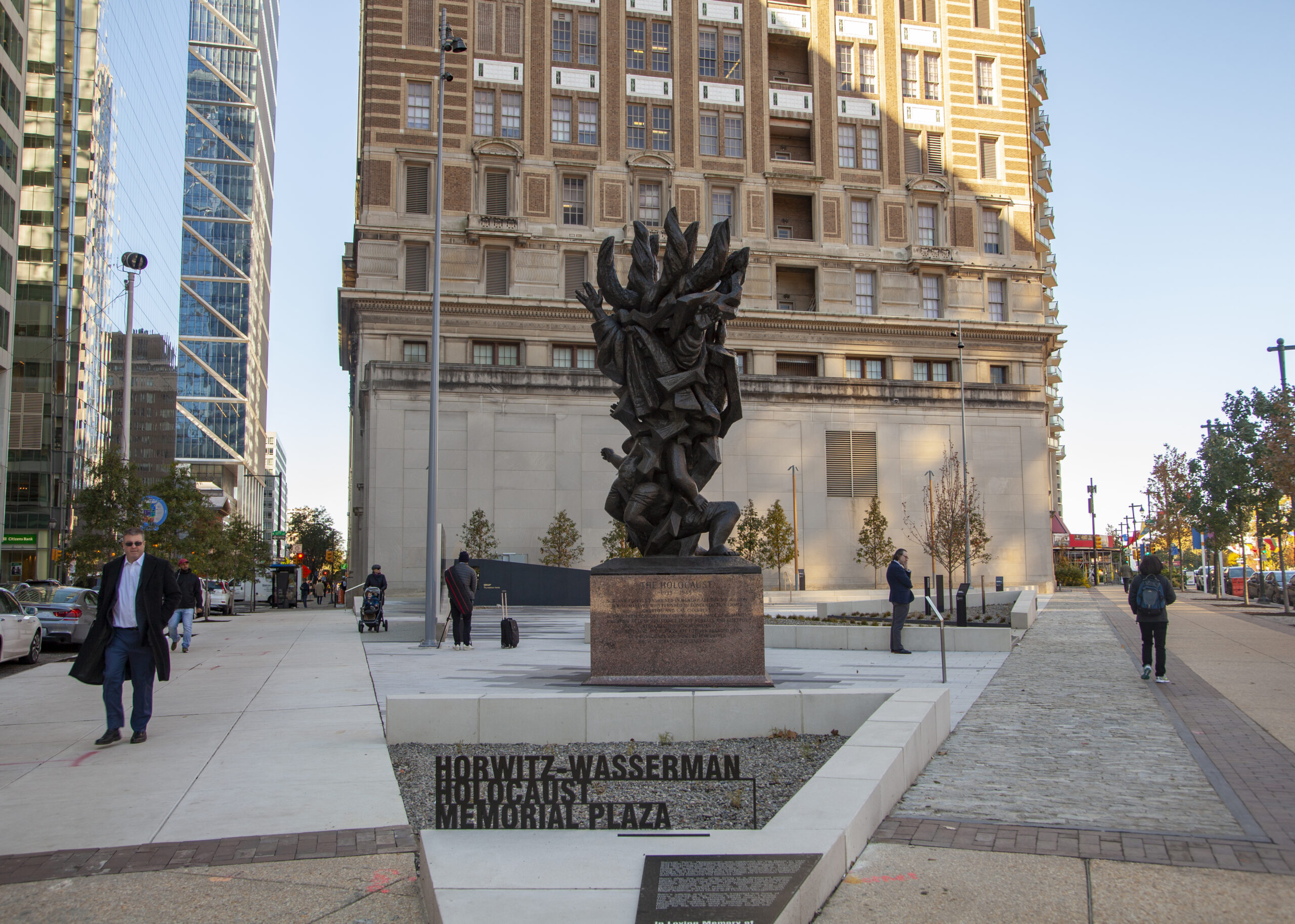 This photo is of the Horwitz-Wasserman Holocaust Memorial Plaza in the afternoon. In the foreground is a concrete marker with the plaza's name. Behind it is the Monument to Six Million Jewish Martyrs with three people milling around. In the background is a small copse of trees and a tan and brown building.