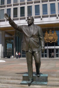 a color photograph of a monument depicting Frank L. Rizzo. The statue is positioned on a set of stairs and the figure of Rizzo is sculpted to appear as if he is walking down the stairs and gesturing towards someone with his right hand. The base of the monument reads "Frank L. Rizzo, Mayor, 1972-1980." Behind the statue is the Municipal Services Building which features the crest of the State of Pennsylvania prominently on the front.