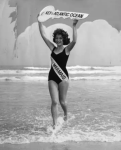 a black and white photograph of a woman standing in ankle-deep water on the beach. She wears a black bathing suit and a sash reading "Miss Mermaid." Above her head she holds a large wooden key that reads "Key to the Atlantic Ocean"
