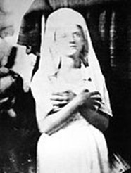 Black and white photograph of a young white woman in a white dress and white veil. Her arms are crossed in front of her as she seems to look past the camera.