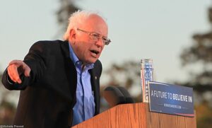 A color photograph of Bernie Sanders speaking at a podium on his presidential campaign in 2016. He faces an unseen audience. His right hand is extended in a pointed gesture. On the front of the podium, a small sign reads "A Future to Believe In. Berniesanders.com"