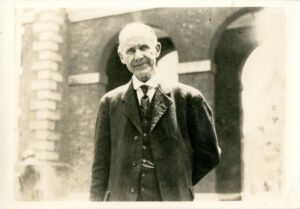 A black and white photograph of Eugene Debs in his elder years, standing with Independence Hall's arcade arches behind him. He is wearing a dark-colored suit and vest and smiles slightly at the camera.