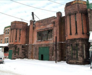 a color photograph of a castle-like building of red stone with green doors. Turrets with narrow vertical slit-like windows stand on either side of the building. It is in a state of decay with boarded and broken windows. Snow covers the sidewalk.