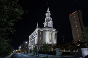 A color photograph showing the Church of Latter Day Saints Temple in Philadelphia. The building is large, white, and features imposing columns on the front facade and a tall steeple topped with a golden statue of a herald angel. The photograph is taken against a night sky emphasizing the illuminated form of the temple.