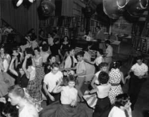 Black and white photograph of a group of teenagers dancing in the center of a room, while the host of the show talks with a young woman in the background.