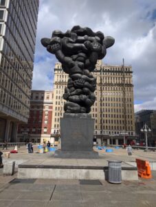 Photograph depicts a large exterior silver metal sculpture made up of twisting limbs stacked upon one another.