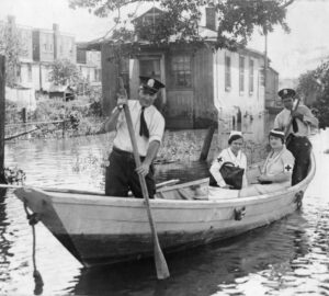 Two uniformed male cops are rowing two uniformed female medical personnel in waters that recently flooded the town of Eastwick.