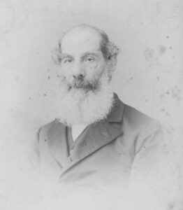 A black-and-white image of a white man with a receding hair line and an impressively full beard. He is wearing a dark coat and waistcoat.