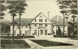 Black-and-white image of a large three-sectioned building with three chimneys, numerous shuttered windows, and a balcony that creates a cover over the main entryway. A large walkway leads up to the building. The gardening is well maintained and trees surround the property.