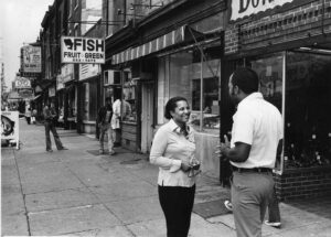 Photograph of two people talking on the sidewalk outside storefronts on Lancaster Avenue